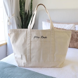 May include: A cream-colored canvas tote bag with a rectangular shape and two handles. The bag has a front pocket and the name "Mrs. Nash" is printed in black script. The bag is sitting on a bed.