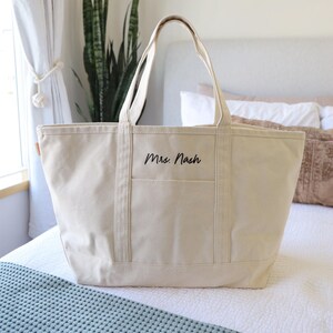 May include: A cream-colored canvas tote bag with a rectangular shape and two handles. The bag has a front pocket and the name "Mrs. Nash" is printed in black script. The bag is sitting on a bed.