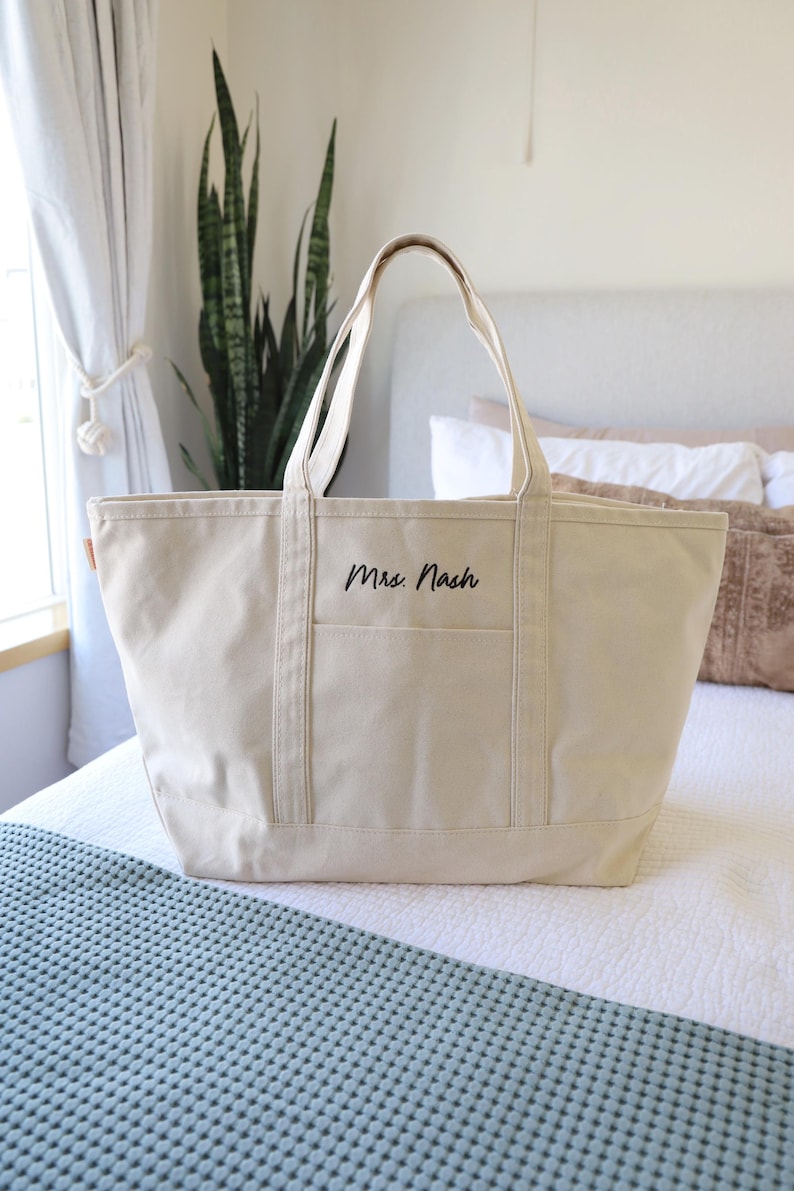 May include: A cream-colored canvas tote bag with a rectangular shape and two handles. The bag has a front pocket and the name "Mrs. Nash" is printed in black script. The bag is sitting on a bed.