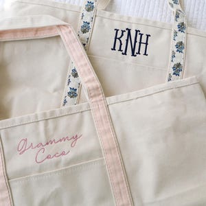 May include: Two off-white canvas tote bags. One has pink trim and the embroidered text "Grammy Coca". The other has blue floral patterned straps and the embroidered initials "KNH".