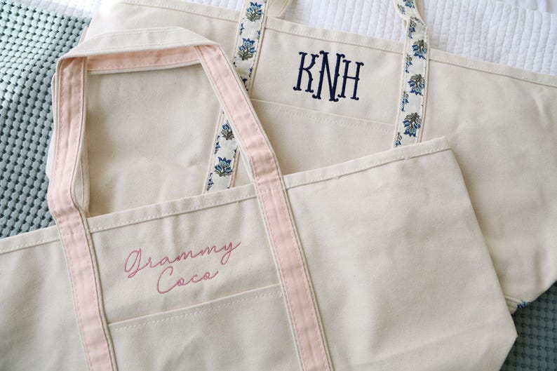 May include: Two off-white canvas tote bags. One has pink trim and the embroidered text "Grammy Coca". The other has blue floral patterned straps and the embroidered initials "KNH".