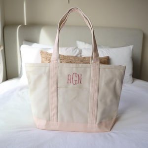 May include: A beige canvas tote bag with pink accents. The bag features a pink bottom and trim, with pink straps. The front of the bag has a pocket and embroidered initials in a reddish-pink color. The bag is sitting on a bed.