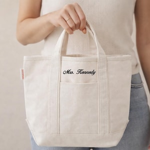 May include: Cream-colored canvas tote bag with a front pocket and two handles. The text "Mrs. Kennedy" is embroidered on the front. A small red tag is visible on the side of the bag.