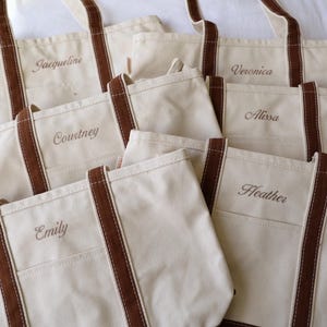 May include: Several off-white canvas tote bags with brown accents and handles. Each bag has a name embroidered on the front: Jacqueline, Courtney, Emily, Veronica, Alissa, and Heather. The bags are arranged on a white surface.