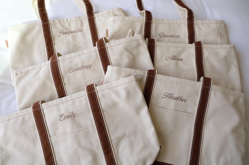 May include: Several off-white canvas tote bags with brown accents and handles. Each bag has a name embroidered on the front: Jacqueline, Courtney, Emily, Veronica, Alissa, and Heather. The bags are arranged on a white surface.