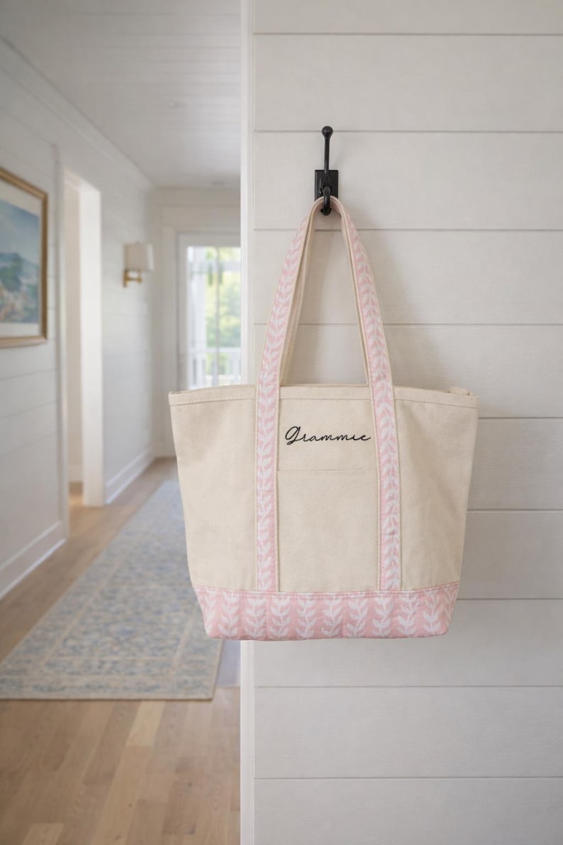 May include: A beige canvas tote bag with pink accents, hanging on a black hook. The bag features pink patterned trim and handles, with the word "Grammie" in black script. The bag is hanging on a white wall.