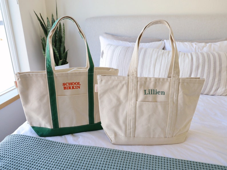 May include: Two canvas tote bags. One is beige with green trim and the text "SCHOOL BIRKIN" in red. The other is beige with green text that says "Lillien". Both have a pocket on the front and sturdy handles.