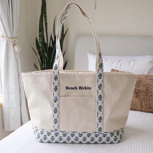 May include: A cream-colored tote bag with blue floral patterned accents on the handles and base. The bag has a front pocket and the words "Beach Birkin" are printed on the front. The bag is sitting on a white bedspread.