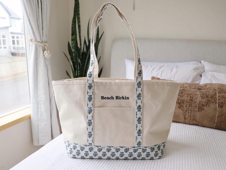 May include: A cream-colored tote bag with blue floral patterned accents on the handles and base. The bag has a front pocket and the words "Beach Birkin" are printed on the front. The bag is sitting on a white bedspread.