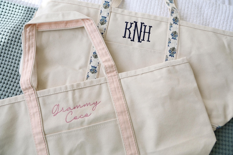 May include: Two off-white canvas tote bags. One has pink trim and the embroidered text "Grammy Coca". The other has blue floral patterned straps and the embroidered initials "KNH".