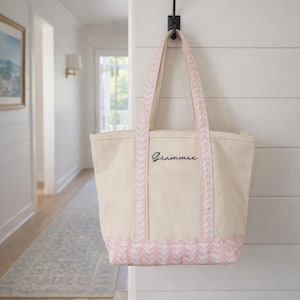 May include: A beige canvas tote bag with pink accents, hanging on a black hook. The bag features pink patterned trim and handles, with the word "Grammie" in black script. The bag is hanging on a white wall.