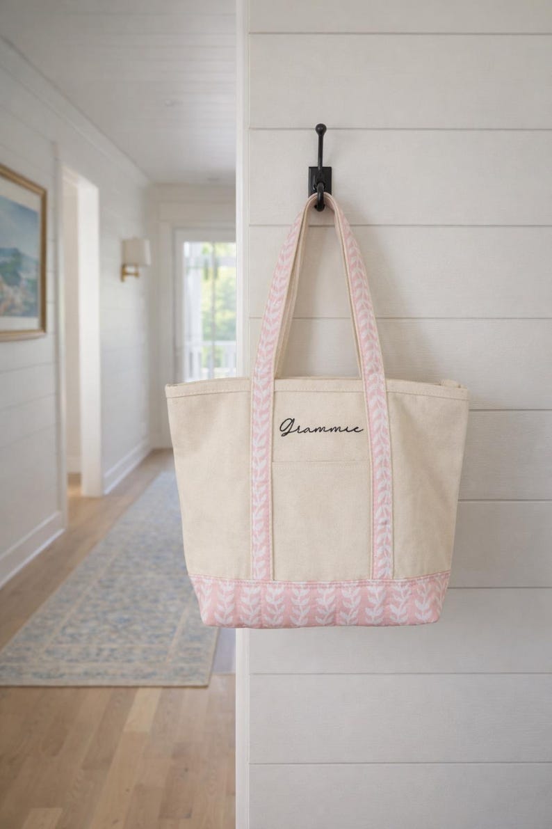 May include: A beige canvas tote bag with pink accents, hanging on a black hook. The bag features pink patterned trim and handles, with the word "Grammie" in black script. The bag is hanging on a white wall.