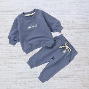 May include: A blue children's outfit, consisting of a long-sleeved sweatshirt and matching sweatpants. The sweatshirt has the name "HENRY" printed on the front. The sweatpants have a drawstring. The outfit is displayed on a light wood surface.
