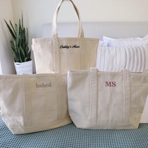 May include: Three canvas tote bags in varying sizes, each with a different embroidered text. The largest bag reads "Oakley's Mom", the medium bag says "baked", and the smallest bag has the initials "MS". The bags are beige.
