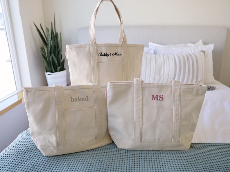 May include: Three canvas tote bags in varying sizes, each with a different embroidered text. The largest bag reads "Oakley's Mom", the medium bag says "baked", and the smallest bag has the initials "MS". The bags are beige.