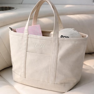 May include: Cream-colored canvas tote bag with a pocket and handles. The bag has the embroidered initials "LH" on the front. Inside the bag are pink items and a white baseball cap with black text. The bag is sitting on a white surface.