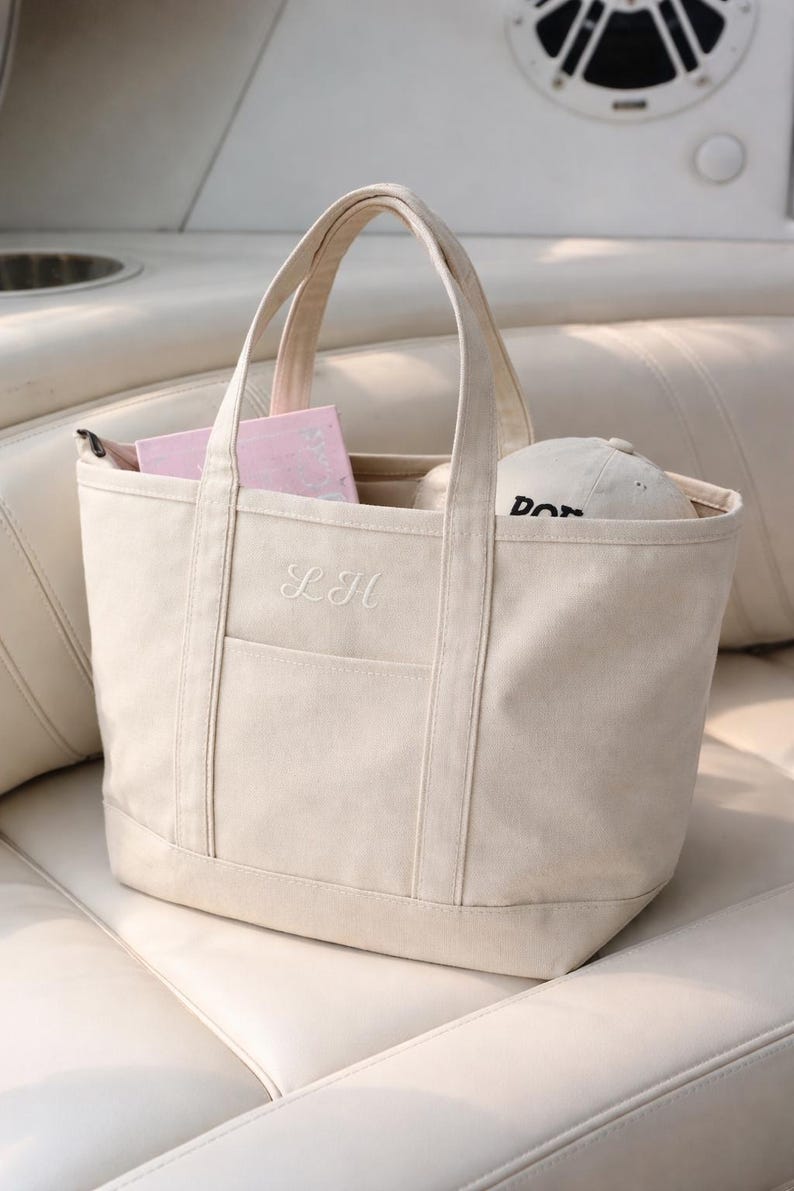 May include: Cream-colored canvas tote bag with a pocket and handles. The bag has the embroidered initials "LH" on the front. Inside the bag are pink items and a white baseball cap with black text. The bag is sitting on a white surface.