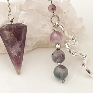 Op de afbeelding: Twee divinatiependels. Eén is een gefacetteerd, paars kristal dat aan een zilveren ketting hangt. De andere heeft een zilveren slangensieraad en paarse en grijze edelsteen kralen. De achtergrond is een wit, kristallijn oppervlak.