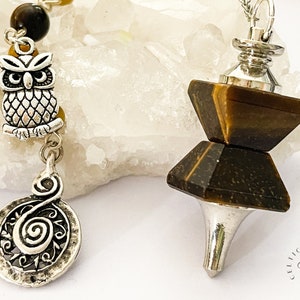 May include: Two pendulums. One has a silver owl charm, a spiral charm, and tiger's eye beads. The other has a tiger's eye crystal with a silver top and a pointed silver base. The background is a white, crystalline surface.