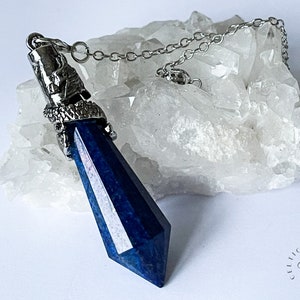 Könnte beinhalten: Ein dunkelblauer Kristallanhänger mit einer silberfarbenen Metallkappe und Kette. Der Kristall ist ein tiefes, facettiertes Blau, und die Kette ist ein zartes Silber. Der Anhänger wird vor einem weißen, strukturierten Hintergrund präsentiert.