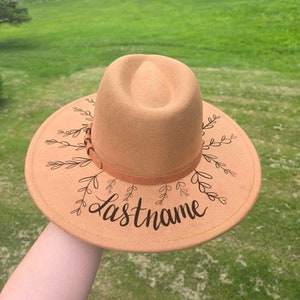 Op de afbeelding: Een beige fedora hoed met een brede rand, versierd met zwarte bladeren en het woord "Lastname". De hoed heeft een bruine band rond de kroon. Een modieus accessoire.
