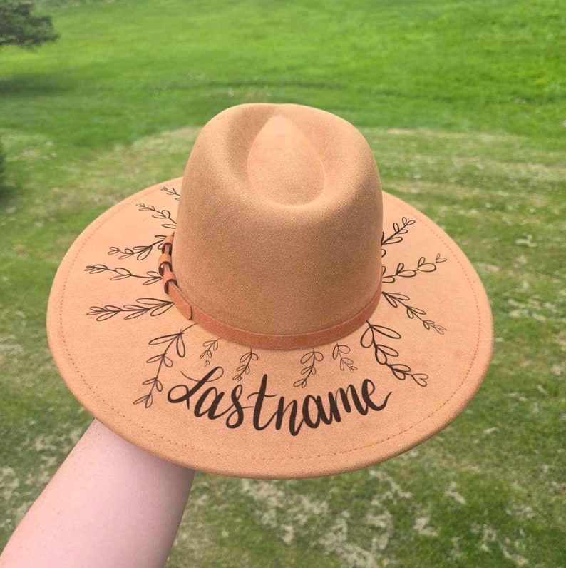 Op de afbeelding: Een beige fedora hoed met een brede rand, versierd met zwarte bladeren en het woord "Lastname". De hoed heeft een bruine band rond de kroon. Een modieus accessoire.