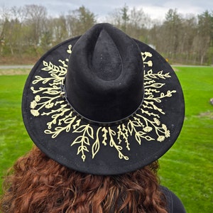 Op de afbeelding: Zwarte fedora hoed met een gouden bloemenpatroon rond de rand. De hoed is gemaakt van een zacht materiaal en heeft een klassieke vorm. Het gouden bloemenpatroon voegt een vleugje elegantie toe. Een stijlvol accessoire.