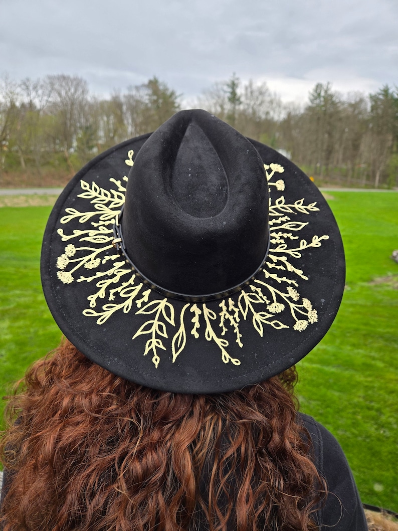 Op de afbeelding: Zwarte fedora hoed met een gouden bloemenpatroon rond de rand. De hoed is gemaakt van een zacht materiaal en heeft een klassieke vorm. Het gouden bloemenpatroon voegt een vleugje elegantie toe. Een stijlvol accessoire.