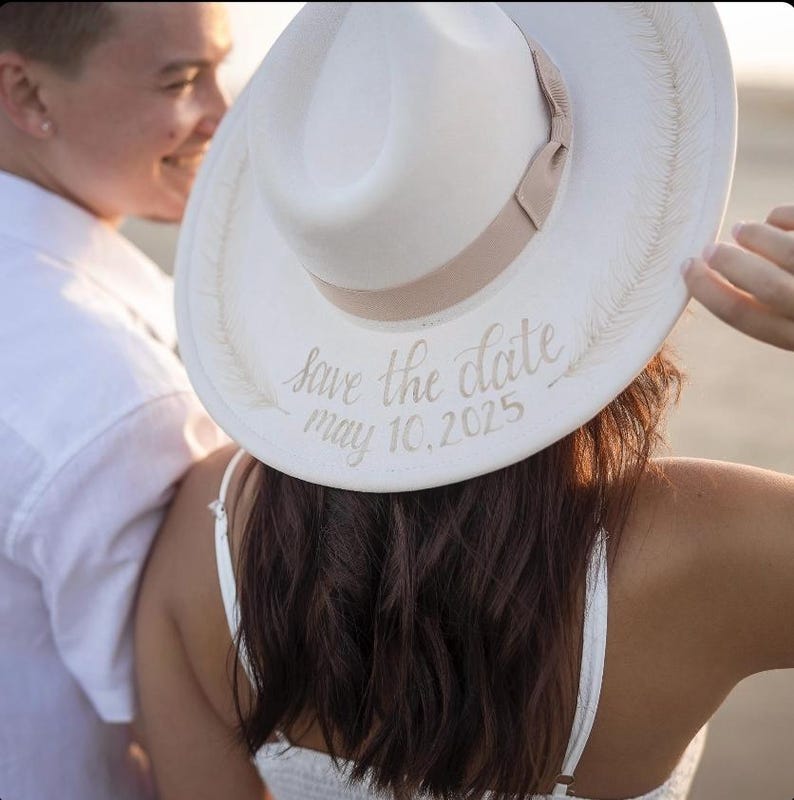 Op de afbeelding: Een witte fedora-hoed met een beige lint en de inscriptie "Save the date May 10, 2025" in elegant schrift. De hoed heeft een brede rand en is versierd met een veermotief. De hoed maakt deel uit van een bruiloftsthema.