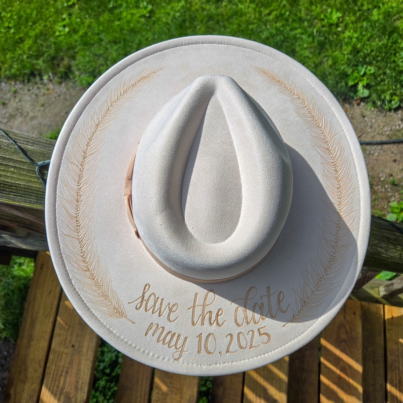 Op de afbeelding: Cr&egrave;mekleurige fedora hoed met een brede rand. De hoed heeft een veermotief en de tekst "Save the date May 10, 2025" in gouden letters. De hoed heeft een bruin bandje rond de kroon.