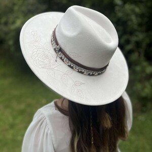Op de afbeelding: Witte fedora-hoed met een decoratieve band en bloemenborduurwerk. De hoed heeft een bruine leren band versierd met strass en een kleine bedel. Een modieus accessoire.