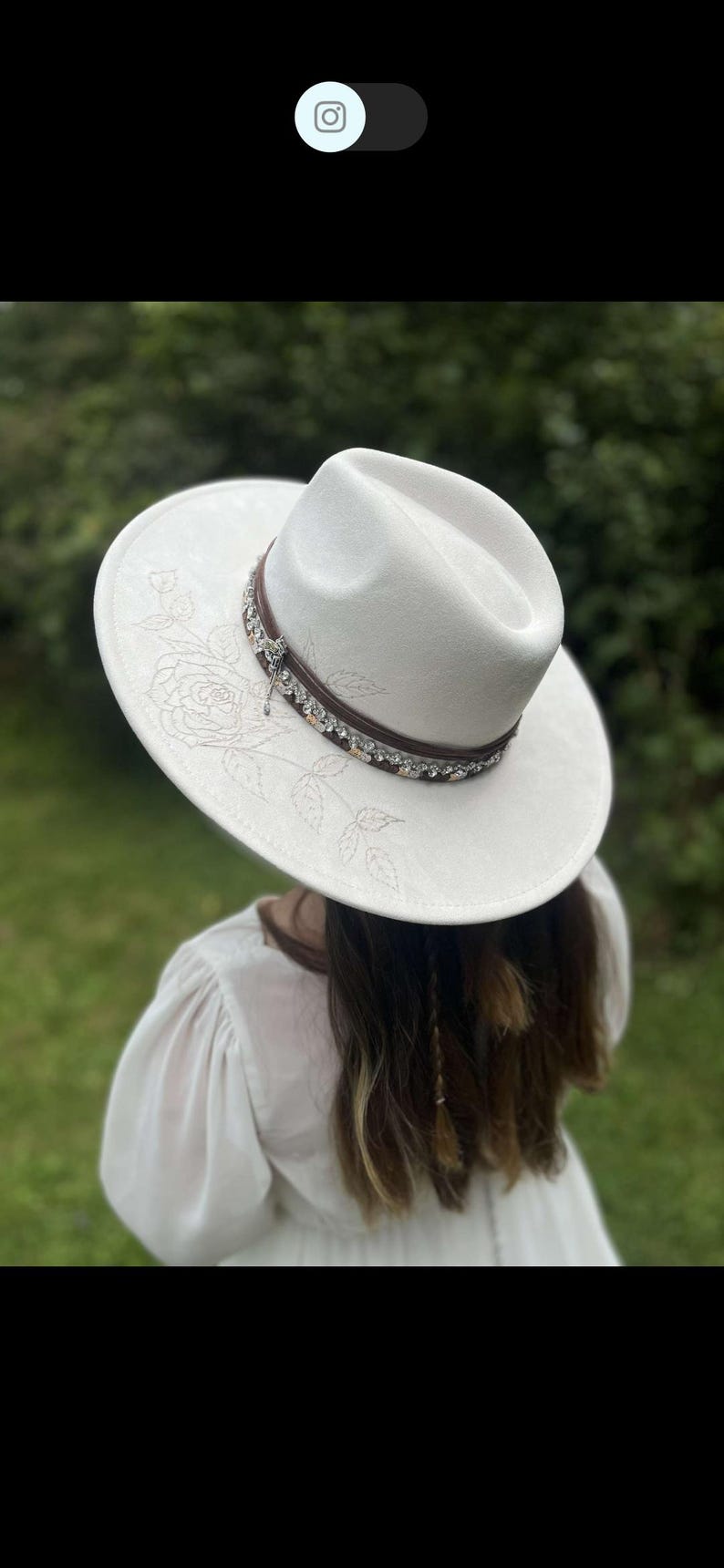 Op de afbeelding: Witte fedora-hoed met een decoratieve band en bloemenborduurwerk. De hoed heeft een bruine leren band versierd met strass en een kleine bedel. Een modieus accessoire.