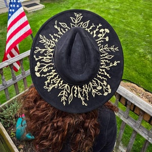 Op de afbeelding: Zwarte fedora hoed met een gouden bloemenontwerp rond de rand. De hoed is gemaakt van een zacht materiaal en heeft een klassieke fedora vorm. Het gouden bloemenontwerp voegt een vleugje elegantie en stijl toe aan de hoed.