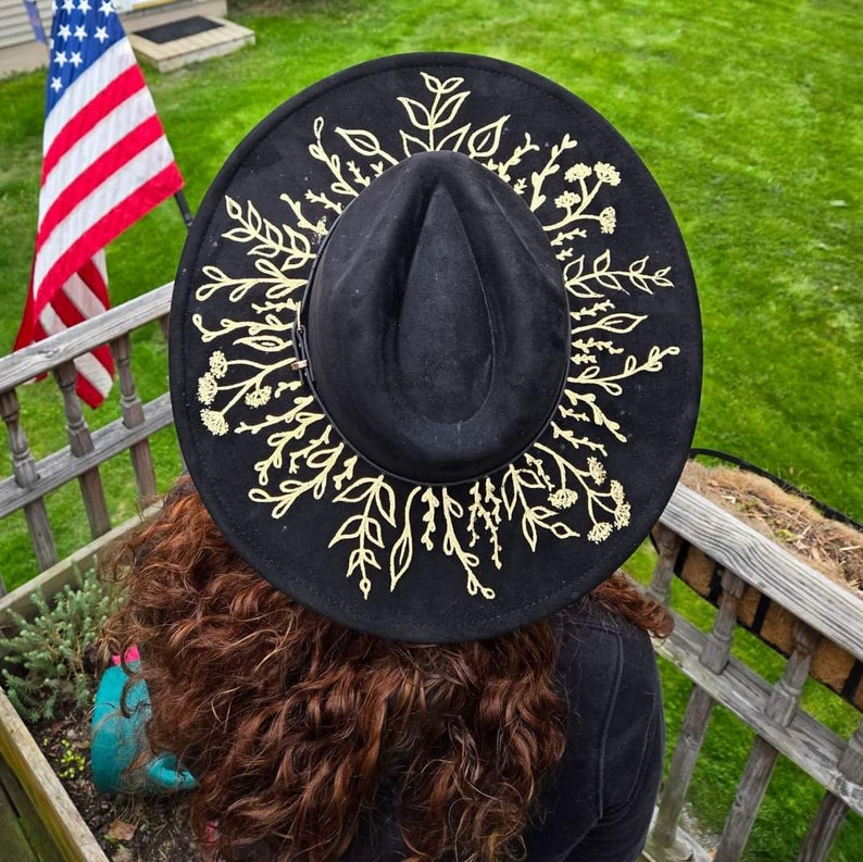 Op de afbeelding: Zwarte fedora hoed met een gouden bloemenontwerp rond de rand. De hoed is gemaakt van een zacht materiaal en heeft een klassieke fedora vorm. Het gouden bloemenontwerp voegt een vleugje elegantie en stijl toe aan de hoed.