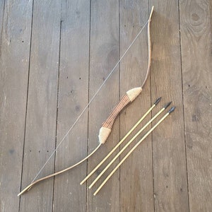May include: A handcrafted wooden bow and three arrows are displayed on a dark wood surface. The bow features a brown handle wrapped with twine and a drawn bowstring. The arrows have black arrowheads.