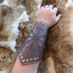 May include: A brown leather arm guard with a black symbol design and silver studs. The arm guard is worn on an arm, with a fur pelt in the background. The arm guard is a Viking-themed accessory.