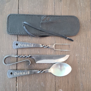 May include: A set of hand-forged metal cutlery including a two-pronged fork, a knife with a looped handle, and a spoon. The fork and spoon feature the name Denni$ engraved on the handle. A black leather storage case with a tie closure is included.