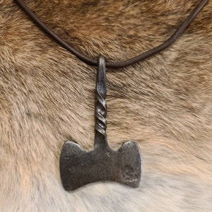 May include: A handcrafted, metal pendant shaped like a hammer, suspended from a brown leather cord. The hammer head is a dark gray color, with a twisted handle. The pendant is displayed against a backdrop of fur.