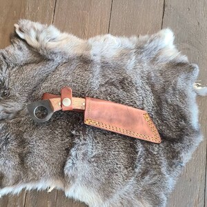 以下が含まれることがあります： 木製の柄と円形の刃が付いた、手作りの小さなナイフ。 ナイフには、革ストラップと金属製のボタンで固定された茶色の革製の鞘が付属しています。 ナイフと鞘は灰色の毛皮の上に置かれています。