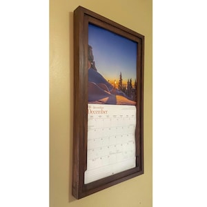 May include: A framed calendar with a wooden border. The calendar displays the month of December, with a scenic image of a snowy mountain landscape at sunset. The frame is a dark brown colour.
