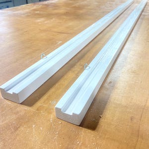 Può includere: Due lunghe barre rettangolari in legno bianco, ciascuna caratterizzata da un canale centrale scanalato che corre lungo l'intera lunghezza del pezzo. Un piccolo gancio a occhiello in metallo argentato è avvitato sul bordo superiore.