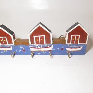 May include: A wooden key holder featuring three red and white painted houses with black roofs. Each house has a white door and sits above a blue sea with a small boat. Gold-colored hooks are below the boats.