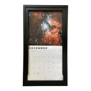 May include: A wall calendar in a black frame featuring a space photograph of a star cluster and nebula at the top. The bottom section displays the month of December with a grid of dates, including the text DECEMBER and small calendar details.