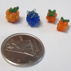 May include: Four miniature glass berry sculptures in orange and blue with green accents. A silver coin is placed next to the berries for size comparison. The berries are clustered and detailed, suggesting a fruit theme.