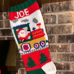 May include: A red, white and green knitted Christmas stocking with the name "JOE" embroidered on the top. The stocking features a Santa Claus in a train with green trees on the bottom.