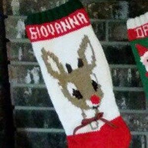 May include: A white Christmas stocking with a red and green trim. The stocking features a knitted reindeer with a red nose and a bell around its neck. The name "Giovanna" is knitted in green letters at the top of the stocking.