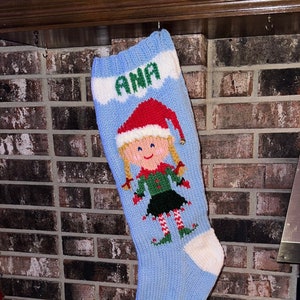 May include: A blue knitted Christmas stocking with a white cuff and a white tip. The stocking features a knitted elf girl with blonde pigtails, a red hat, and a green and black outfit. The stocking is personalised with the name "ANA" in green letters.