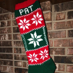 May include: A red and green knitted Christmas stocking with a white snowflake pattern. The stocking has the name "PAT" embroidered on the top.