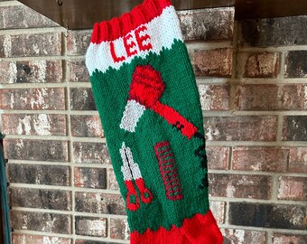May include: A red and green knitted Christmas stocking with the name "LEE" embroidered on the top. The stocking is decorated with a red axe, scissors, and a comb.