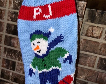 May include: A blue knitted Christmas stocking featuring a snowman skiing down a red and white mountain. The stocking has a red band with the name "PJ" knitted in white.